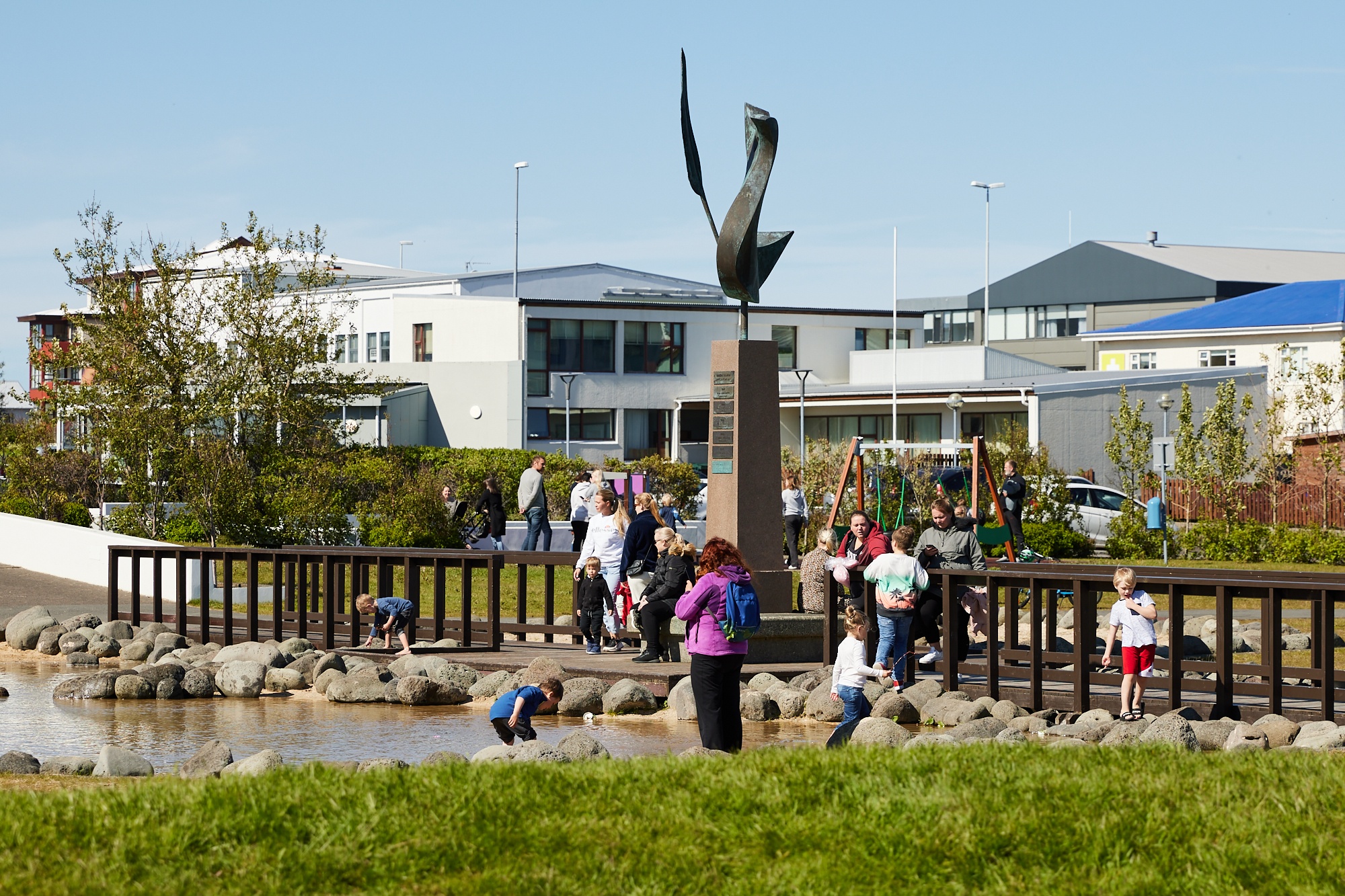 Families and children gather around a small shoreline and wooden bridge near a modern sculpture in a community park in Reykjanesbær, Iceland, reflecting the city’s dynamic and family-friendly environment.
