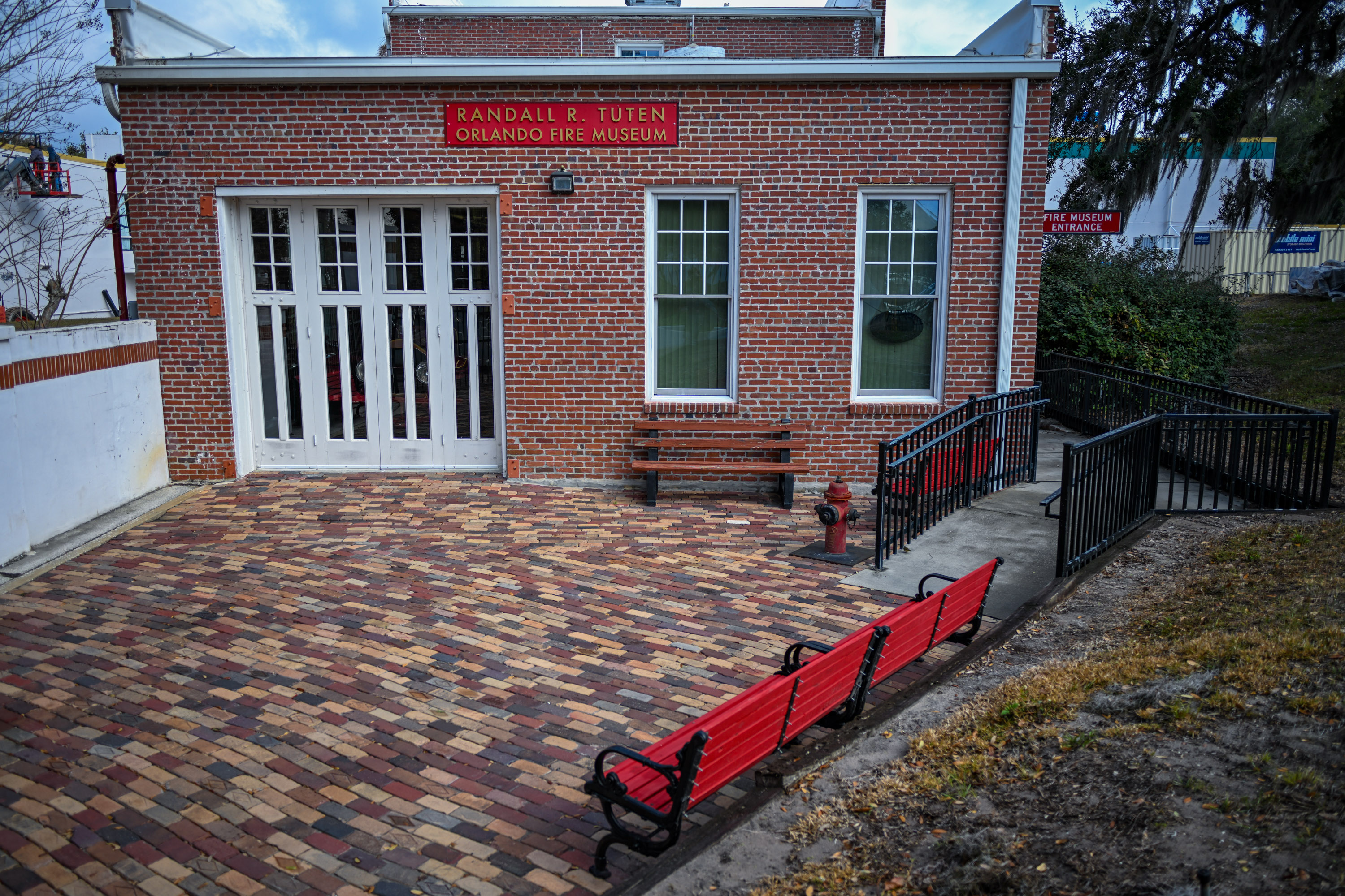 The entrance of the Orlando Fire Museum with red and brown benches in the front. The right side of the building has a fenced pathway which is where the entrance is.