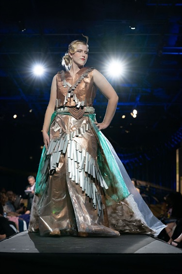 Ellie Magray models a design by Debra Wilson and Heather Magray. The model poses in a copper and teal-colored gown made from construction waste like office blinds and thin plastic packaging.