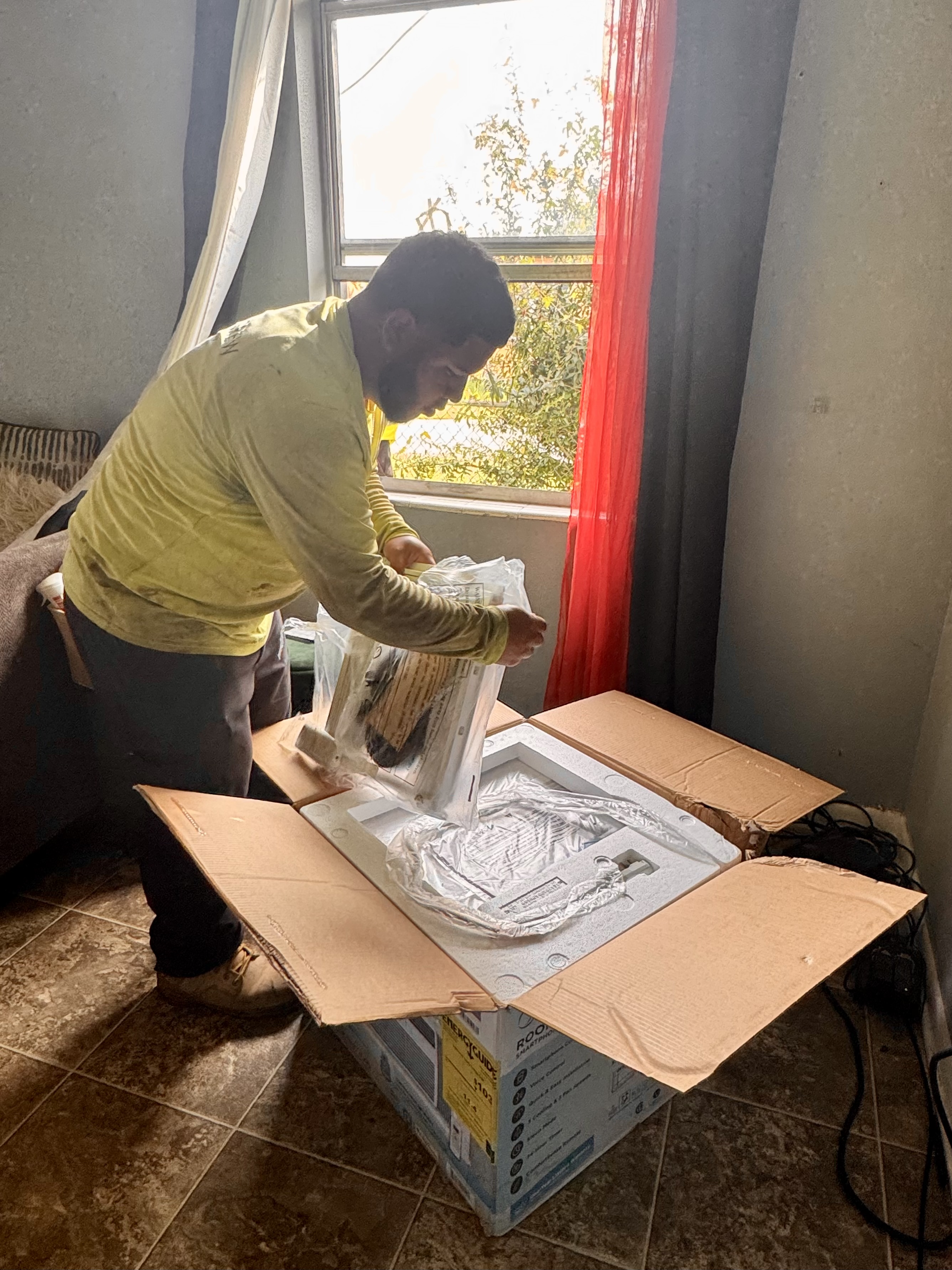 A resident opening a box with a portable AC unit in a room. The window is open and has red curtains.