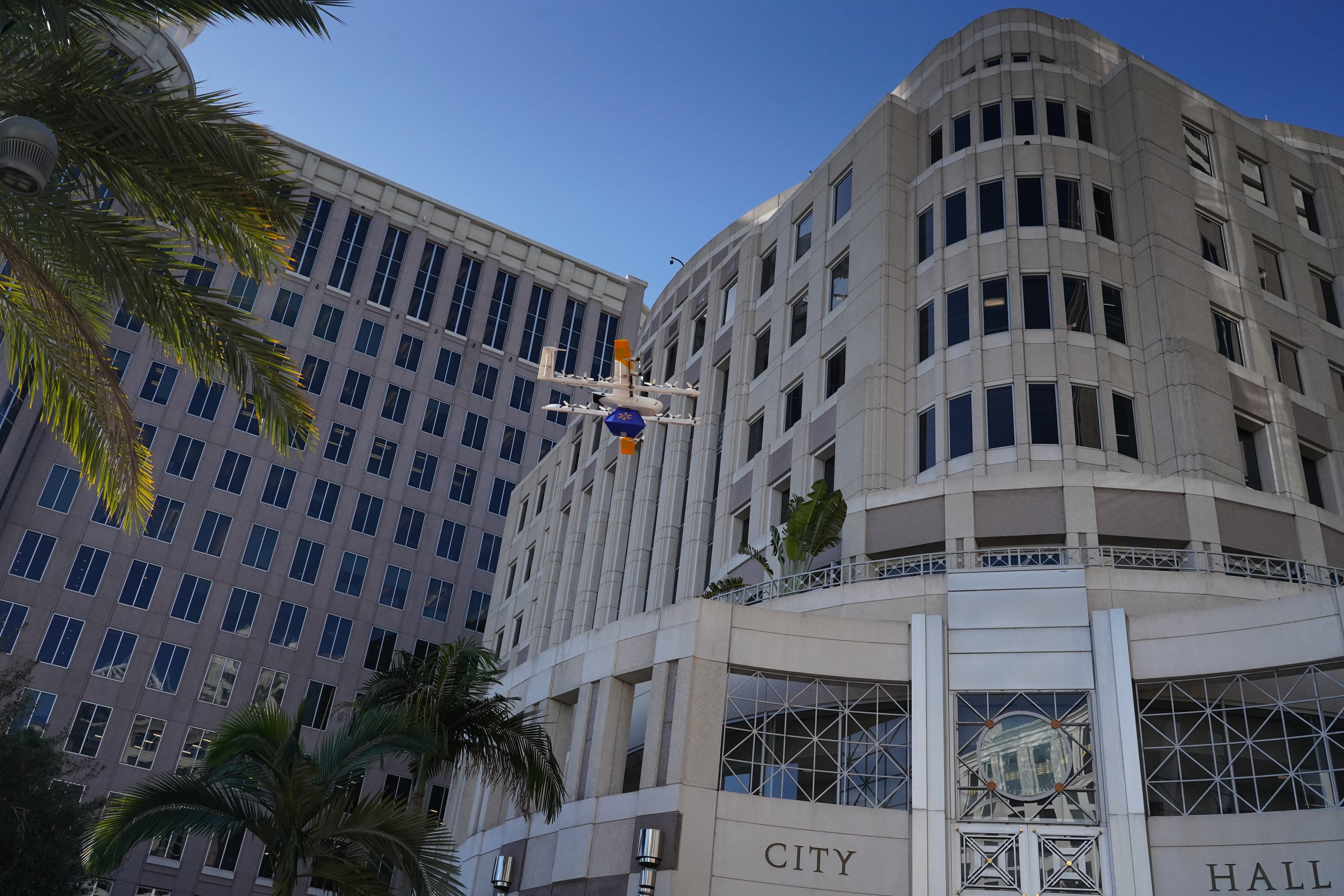 A small delivery drone flies in front of Orlando City Hall with tall windows and palm trees under a bright blue sky.
