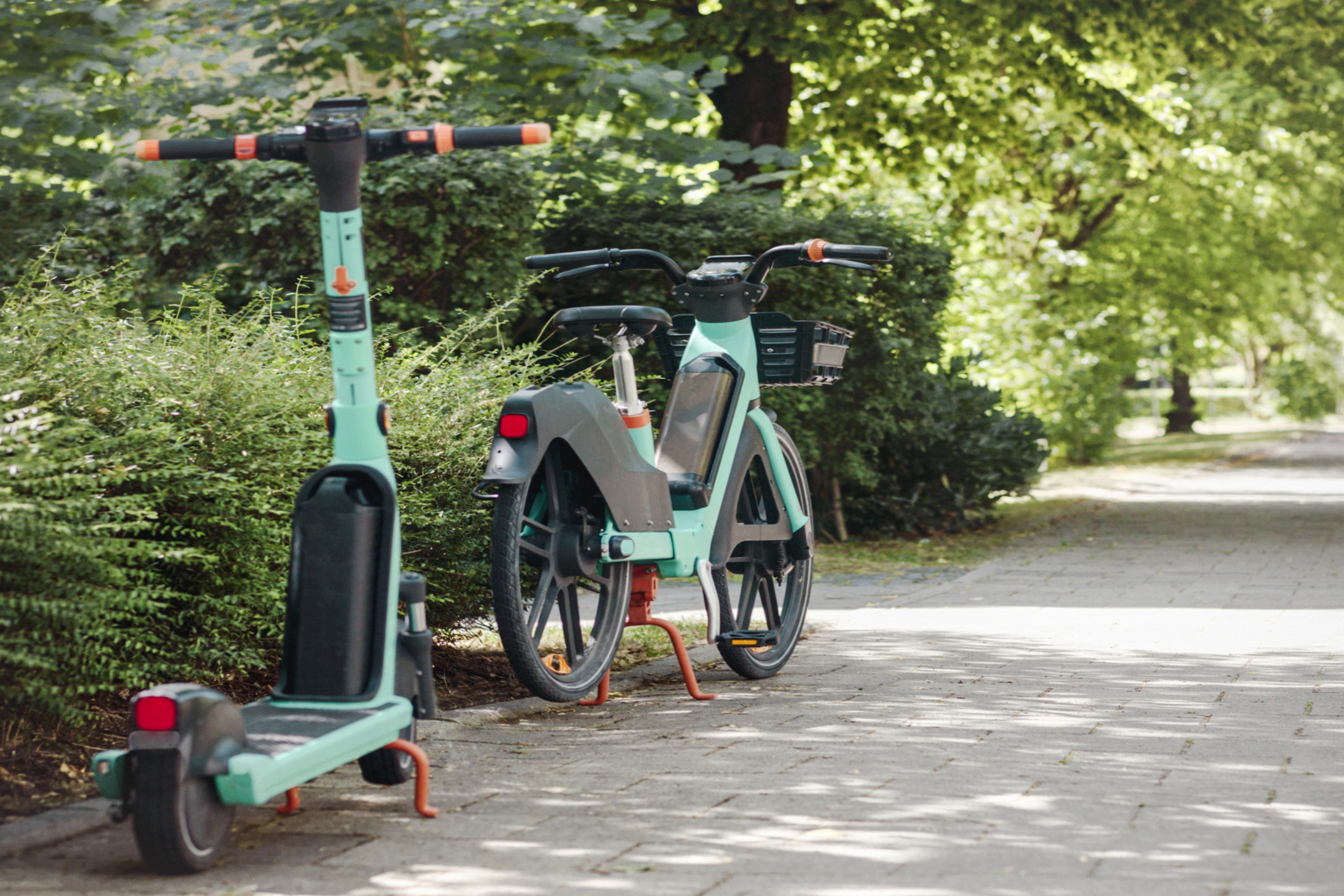 A teal e-scooter and a teal e-bike parked along a walkway. There are bushes and trees along the walkway.