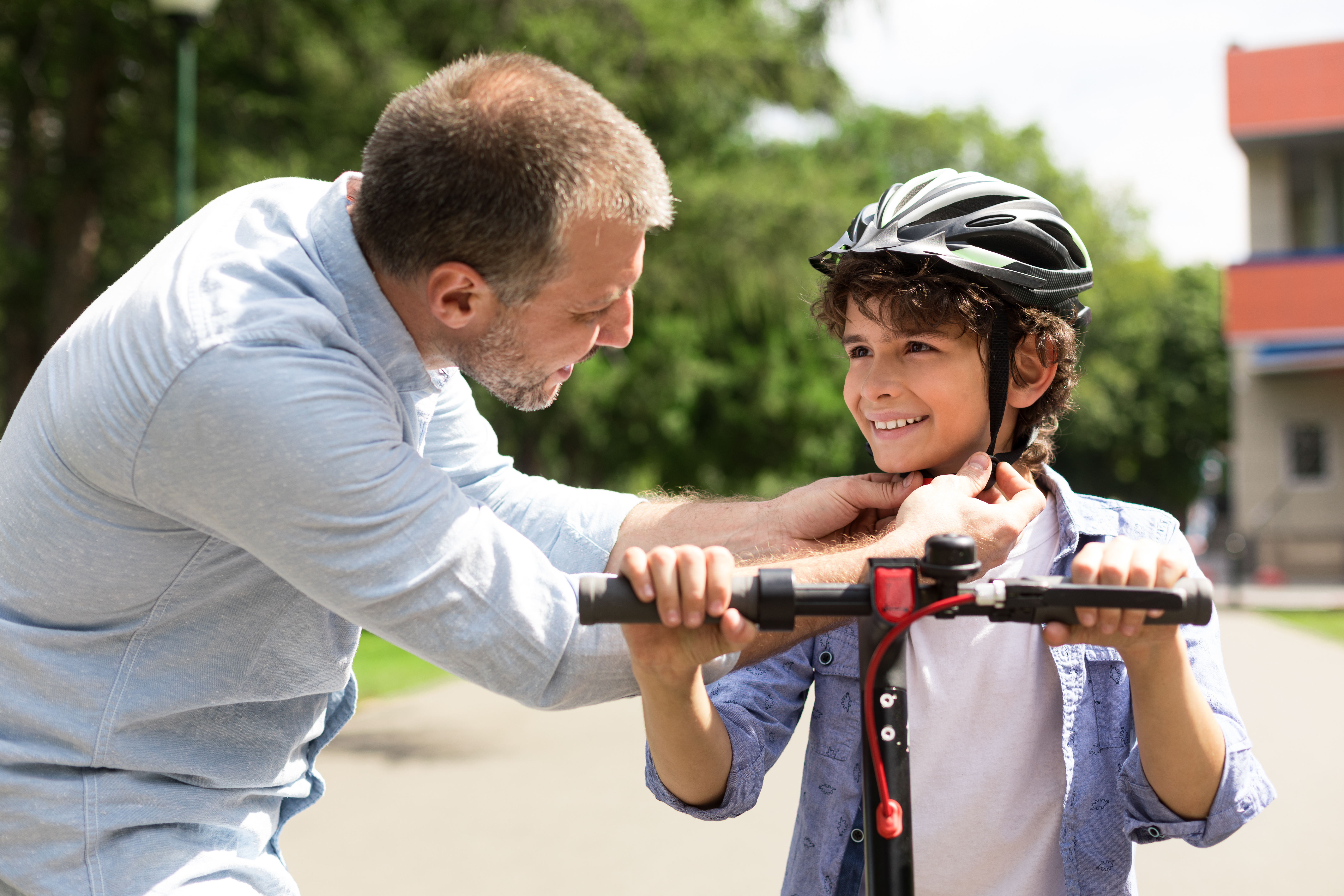 An adult adjusts the chin strap of a safety helmet on a child who is holding the handlebars of a scooter in an outdoor park setting. Trees and a building are visible in the background.