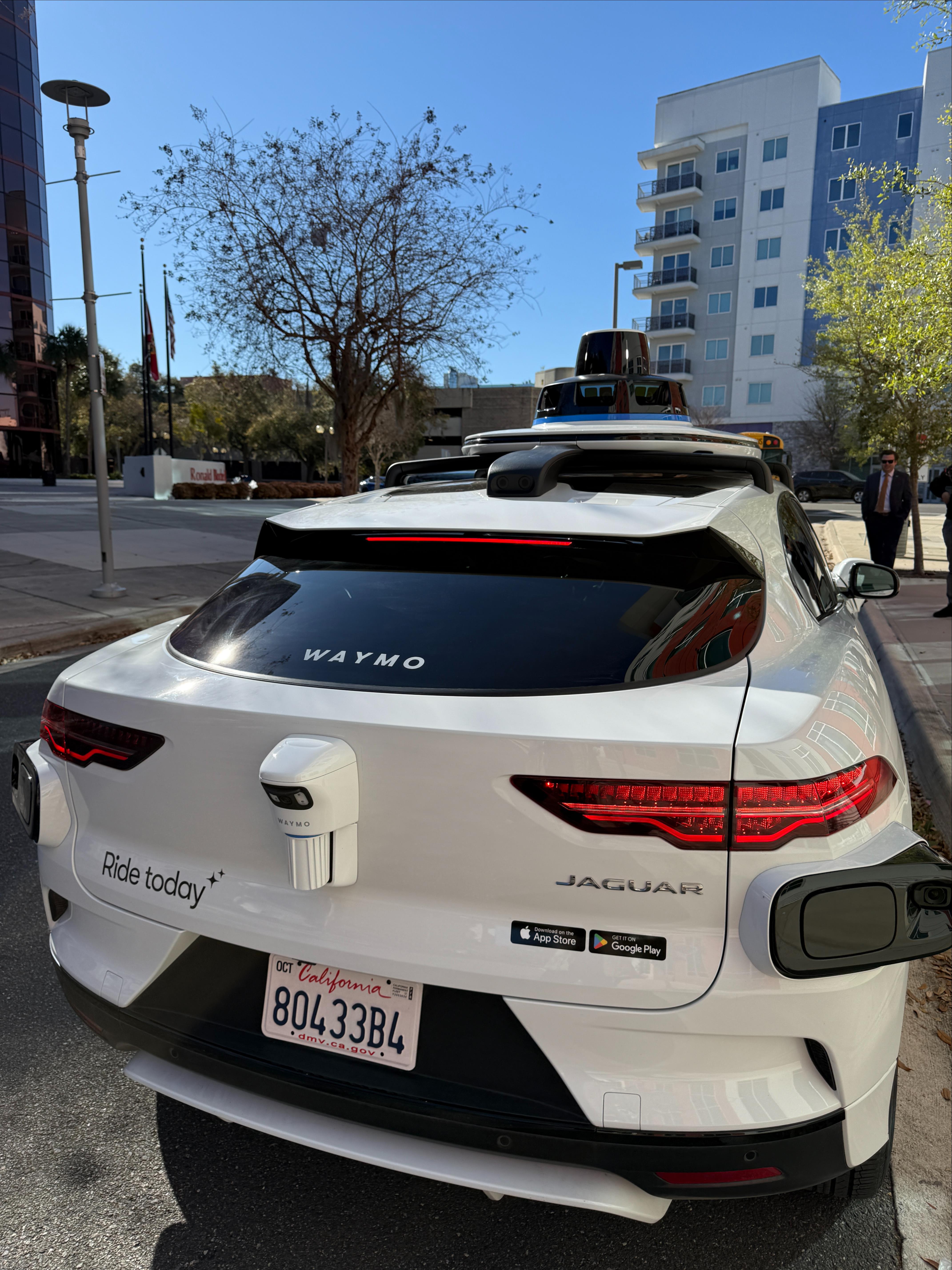 A white Waymo Jaguar autonomous vehicle parked on a city street, showing its rear sensors, with buildings and trees in the background on a sunny day.