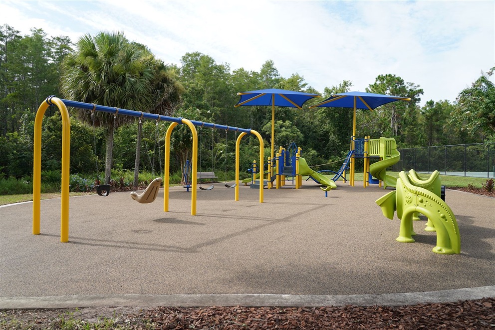 A bright outdoor playground with yellow and blue swings, green slides, large blue shade canopies, and a soft rubber surface, set beside dense trees and greenery.
