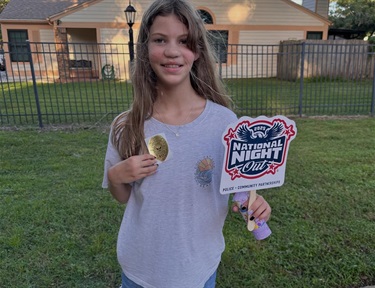 A young girl holding a police badge sticker in one hand and a sign in her other hand that reads national night out she is standing outside with a fence and house behind her