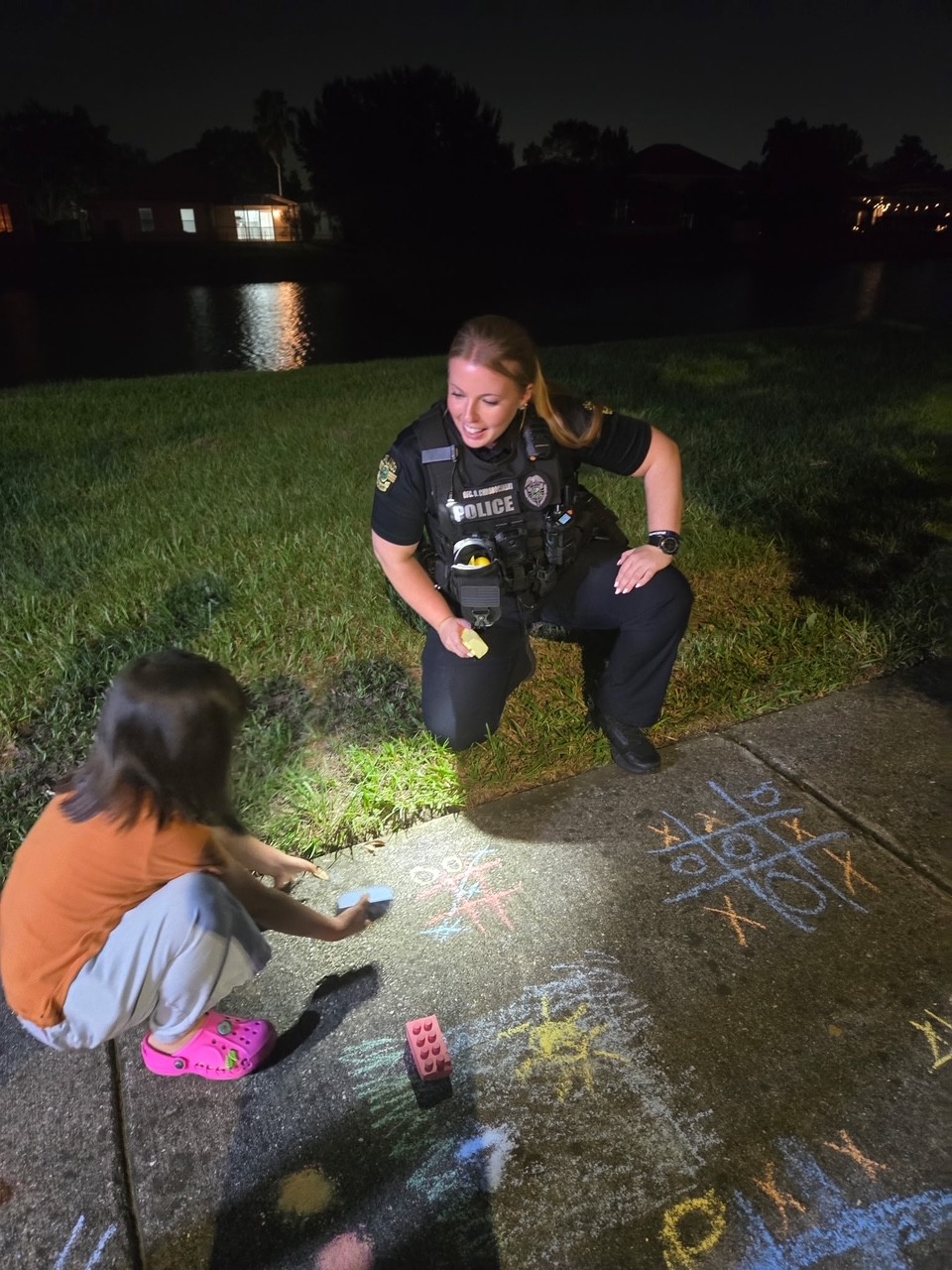 A Police Officer taking a knee in the grass to talk to a little girl making sidewalk art using chalk it is dark out and her chalk art and the officer can be seen with a flashlight