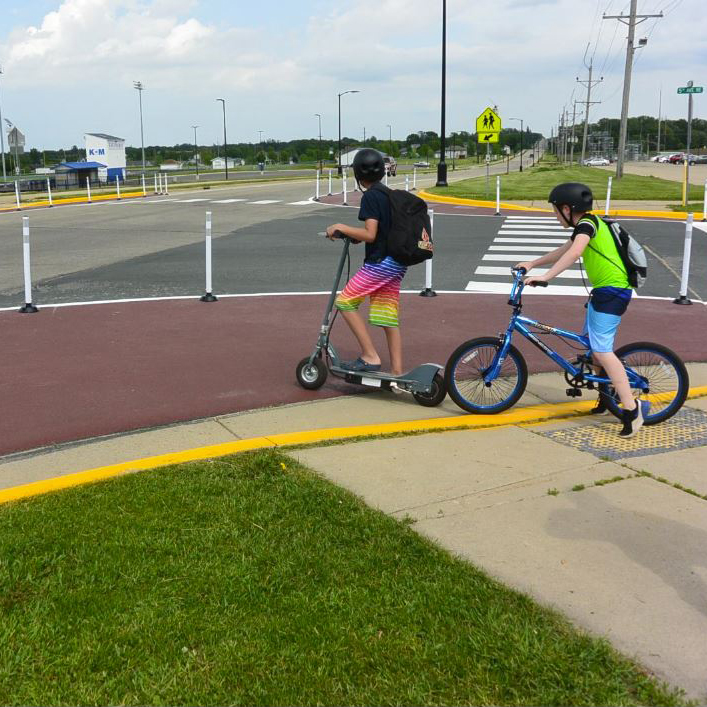 Two youth on a scooter and bike prepare to cross an intersection which is protected with a wide bulb out. The bulb out is lined with flexible posts and the asphalt is a dark red color to increase visibility
