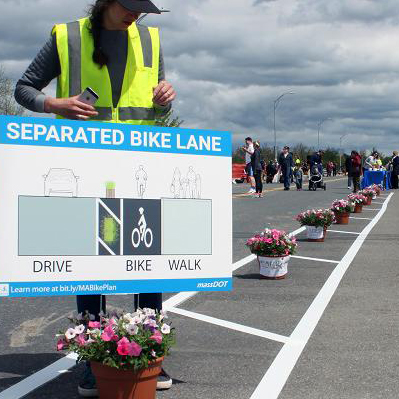 A shoulder repurposed for people biking. Striping on the ground marks a buffer between the temporary pathway and the vehicle lane. A person is placing a sign that says "Separated Bike Lane" in a planter within the painted buffer.