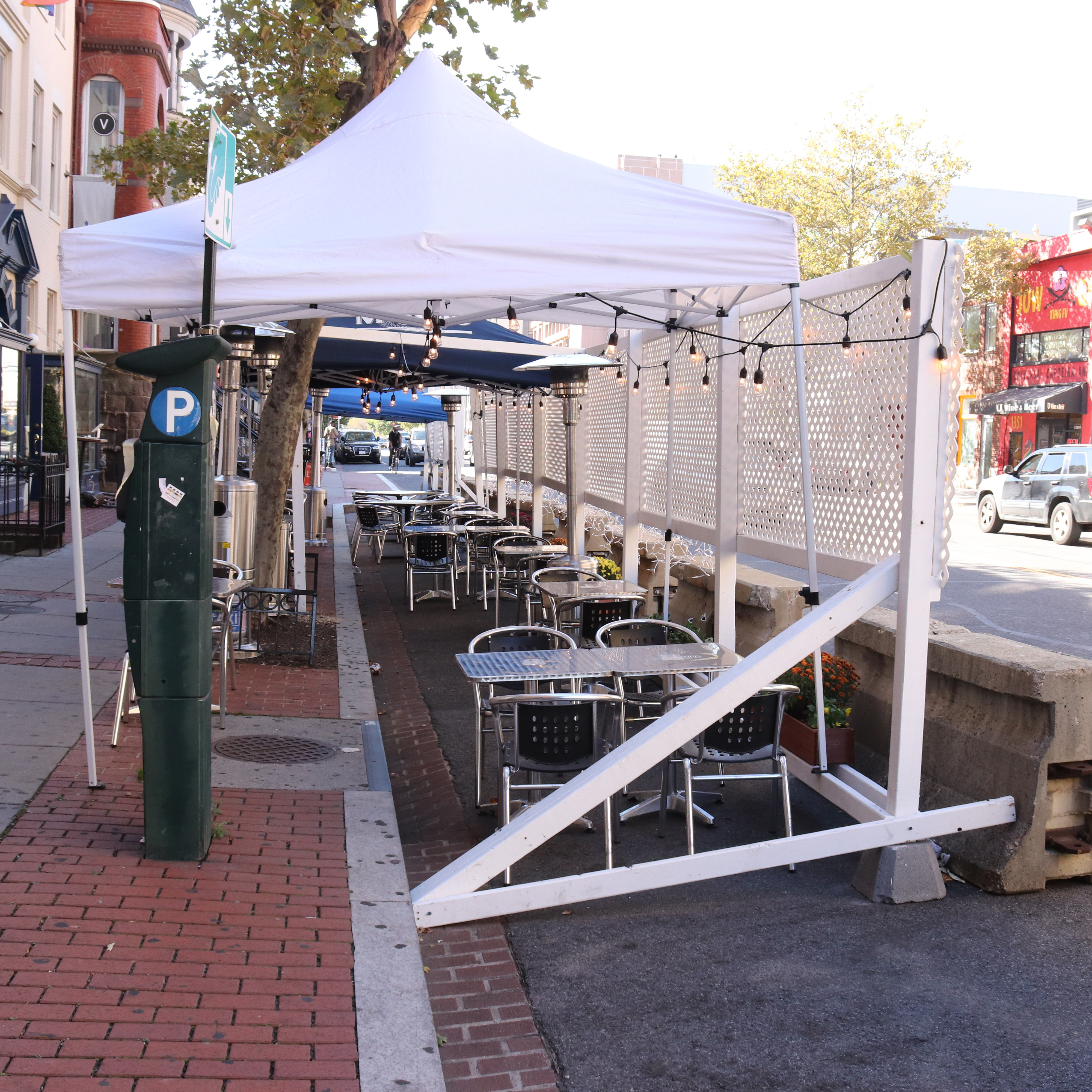 A photograph of an outdoor dining area built within what was once an on-street parking lane. Pop-up tents provide shade to diners.
