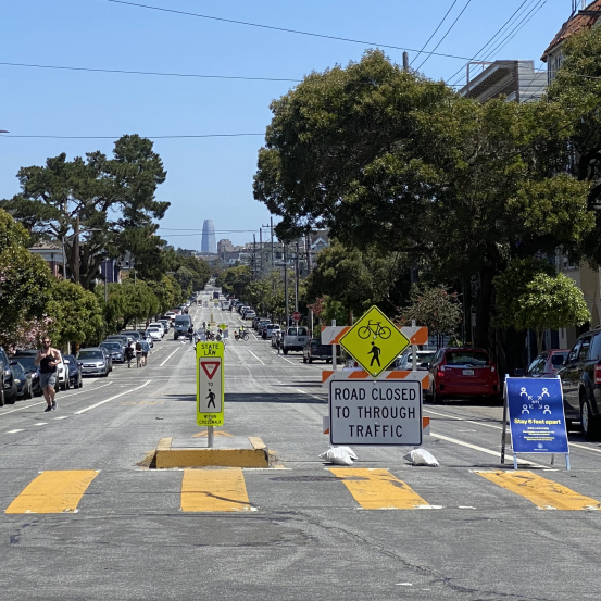 Traffic signs, including a Type III Barricade, close a roadway to through traffic to reserve space for people walking and biking.