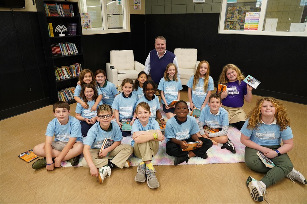 Mayor Dyer and several children sitting on the ground in a reading corner. The children are wearing Mayor Buddy's Book Club shirts and some are holding books